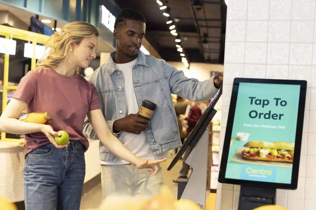 Two customers at a Centra self-order kiosk, with a 'Tap To Order' screen, featuring a woman holding a green apple and a man with a coffee cup, preparing to use the digital ordering system