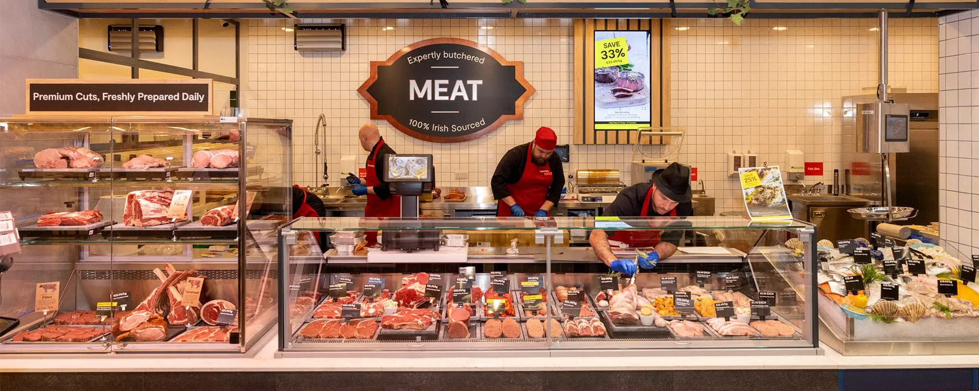 SuperValu meat counter with butchers in red aprons, displaying a wide range of fresh 100% Irish sourced meats in refrigerated display cases, with 'Premium Cuts, Freshly Prepared Daily' signage