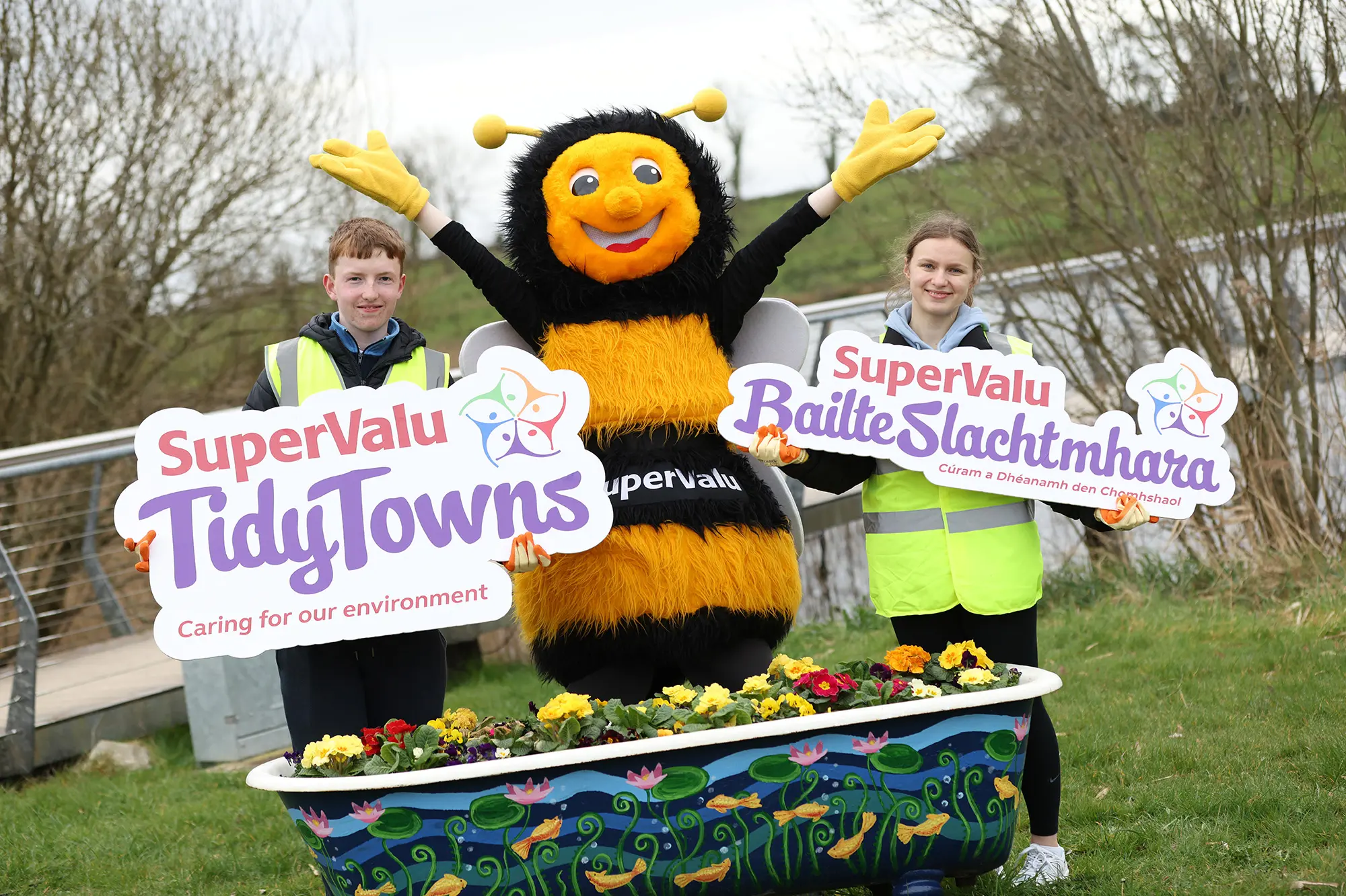 A smiling bee mascot stands with arms raised between two young volunteers wearing high-visibility vests. They hold SuperValu TidyTowns and SuperValu Baile Slachtmhara signs behind a colourful bathtub planter filled with flowers in a grassy outdoor setting.