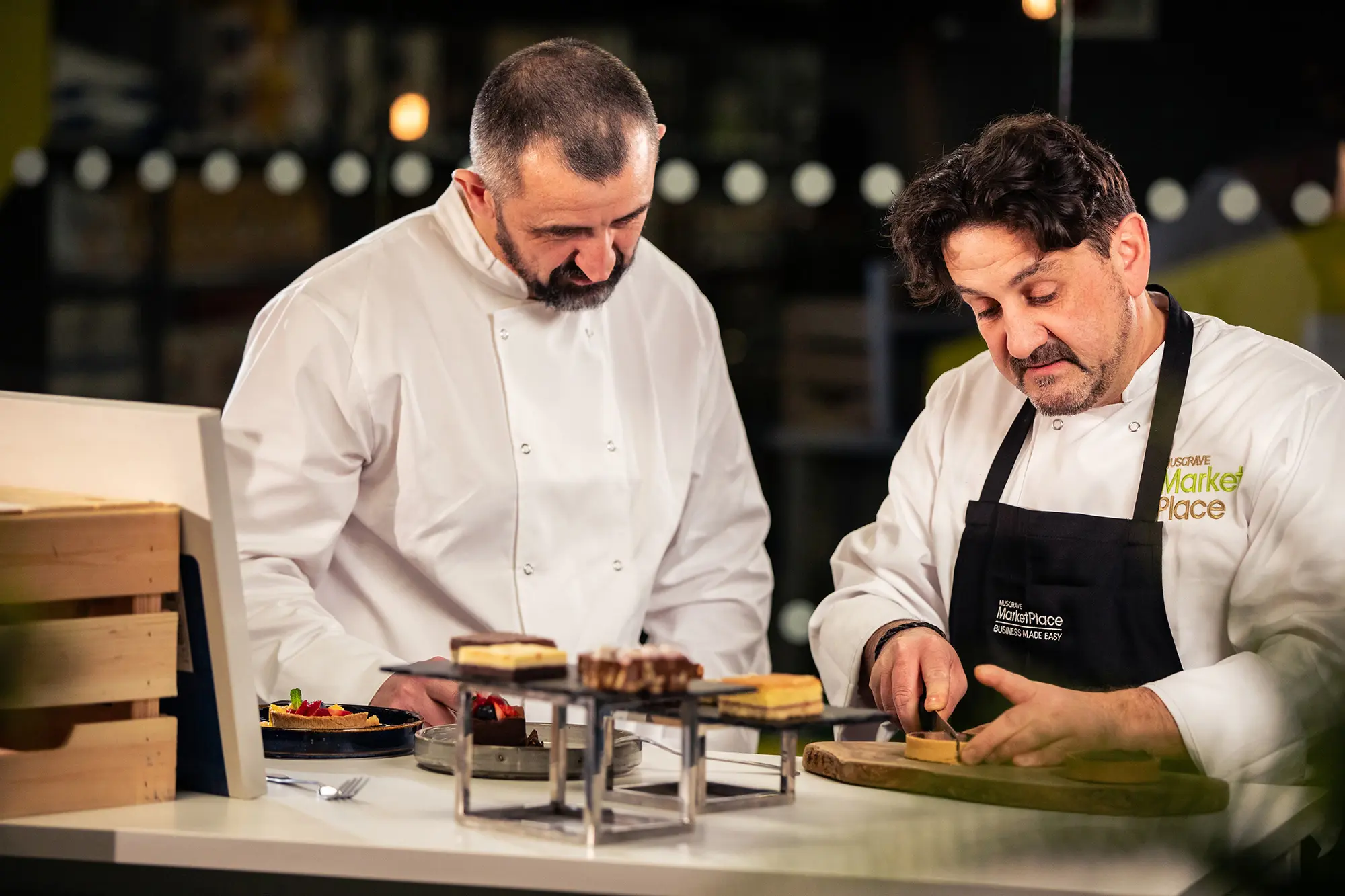 Two chefs prepare and plate desserts at a counter during a food demonstration, with one chef carefully slicing a pastry while the other looks on. The chef's apron features the Musgrave MarketPlace logo.