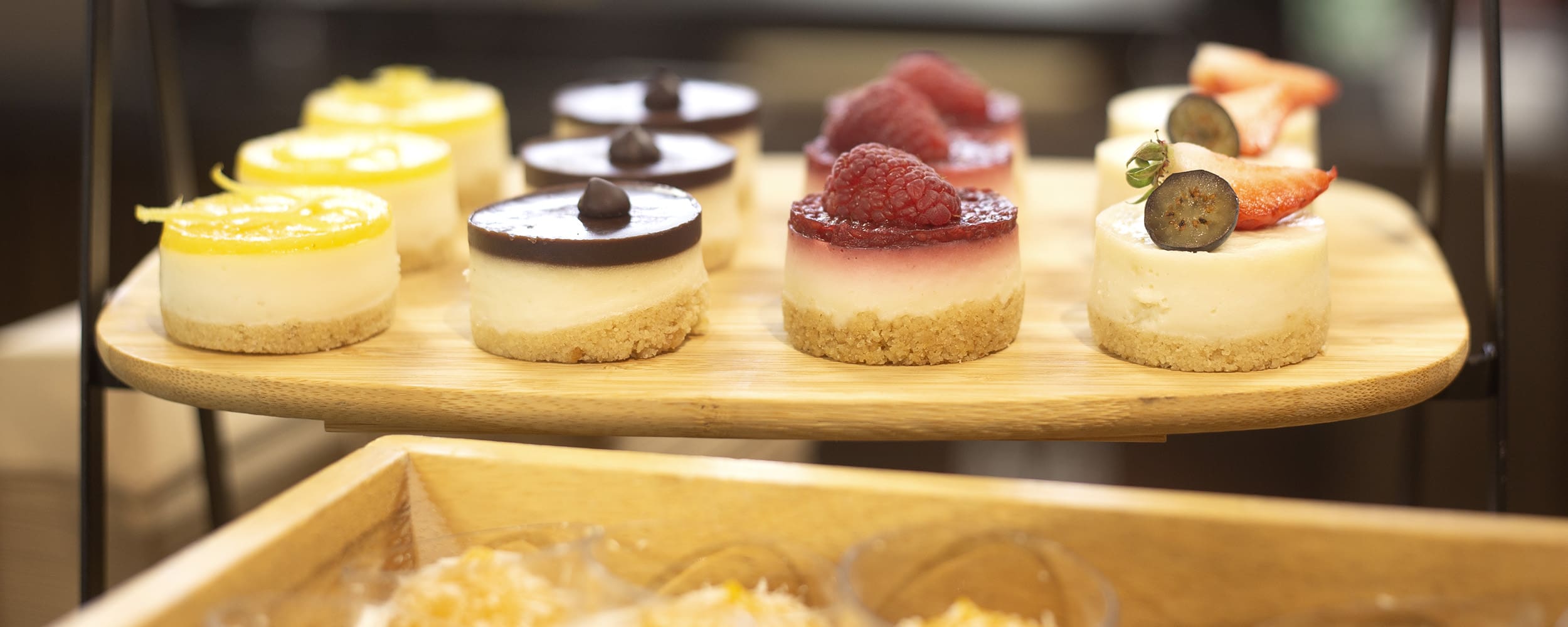 Assorted mini cheesecakes on a wooden platter, featuring flavours including lemon, chocolate, raspberry, and fruit-topped varieties with fresh berries and sliced fruits