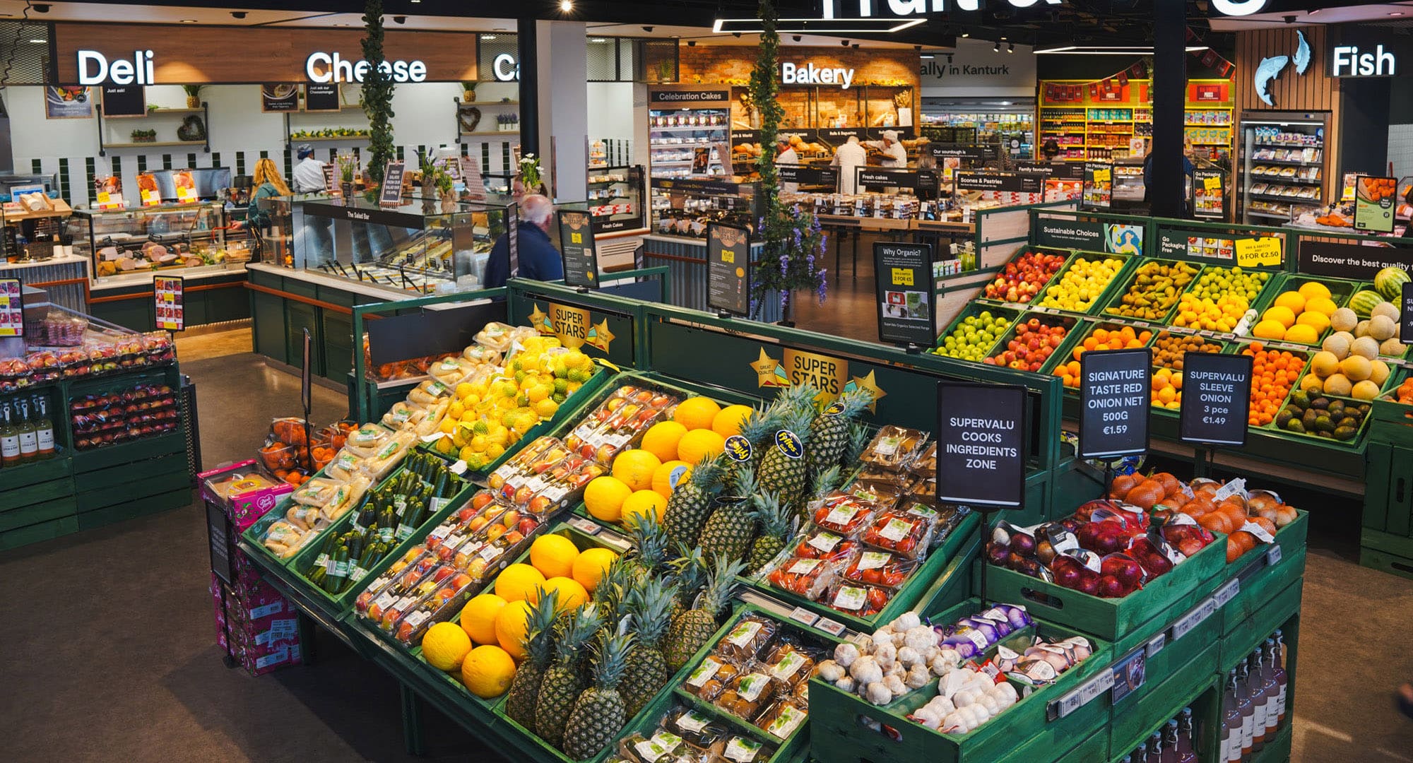 SuperValu grocery store produce section with colourful displays of fresh fruits and packaged produce, with deli, cheese, fish and bakery sections in the background