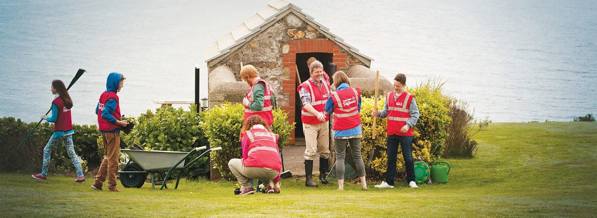 "SuperValu TidyTowns volunteers in bright pink safety vests working together on a community landscaping project, with gardening tools and a wheelbarrow near a stone building overlooking a grassy coastal area